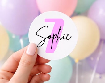 May include: A white circular sticker with the name "Sophie" in black script and a pink number 7. The sticker is held in front of a backdrop of pastel-colored balloons, including pink, purple, and yellow.