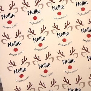 May include: A sheet of adhesive labels featuring a repeating reindeer graphic with brown antlers, beige ears, and a red nose. Each label includes the name Nellie printed in dark blue text and the phrase Love from Santa x below it.
