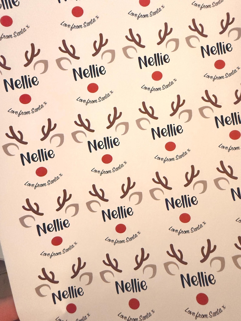 May include: A sheet of adhesive labels featuring a repeating reindeer graphic with brown antlers, beige ears, and a red nose. Each label includes the name Nellie printed in dark blue text and the phrase Love from Santa x below it.