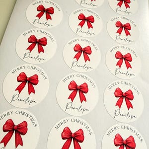 May include: A sheet of fourteen circular white stickers, each featuring a red ribbon bow illustration, the text Merry Christmas in an arched font, and the name Penelope written in a black cursive script below the bow.