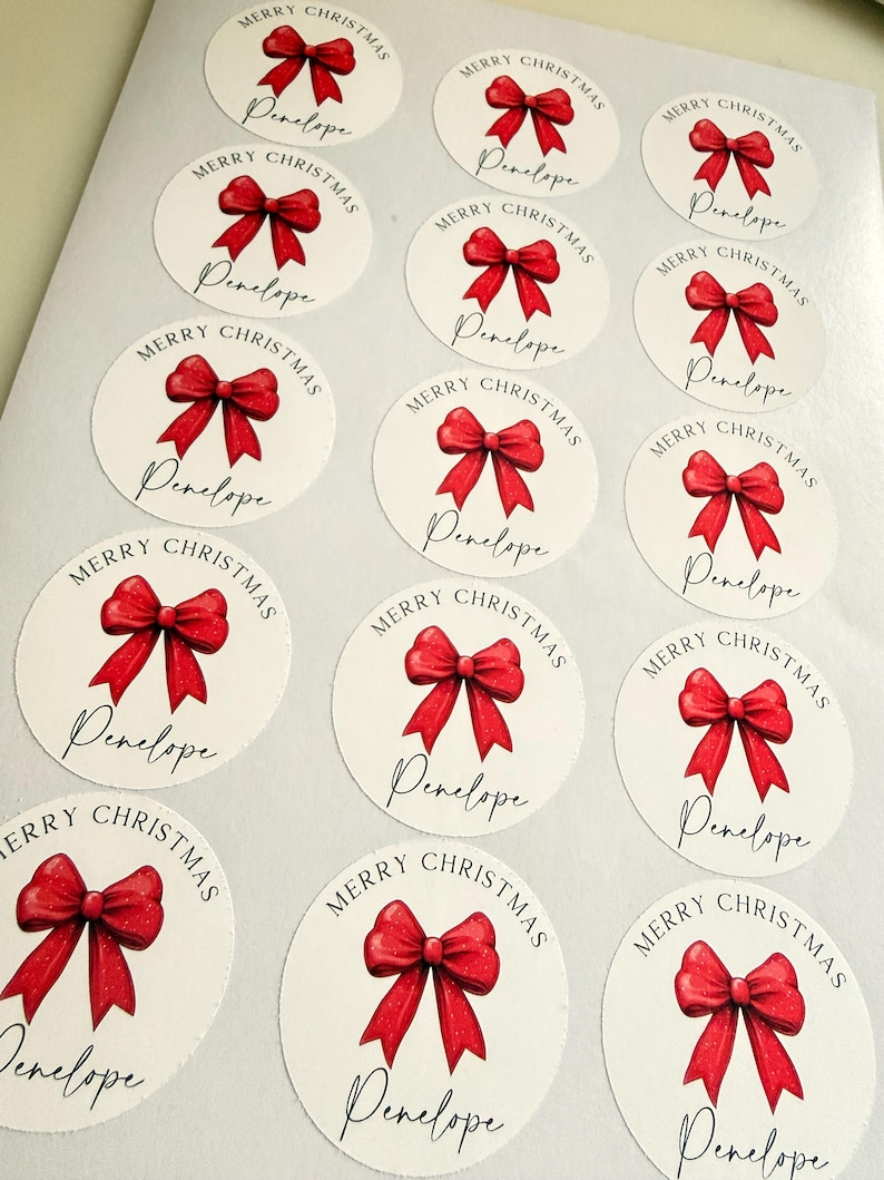 May include: A sheet of fourteen circular white stickers, each featuring a red ribbon bow illustration, the text Merry Christmas in an arched font, and the name Penelope written in a black cursive script below the bow.