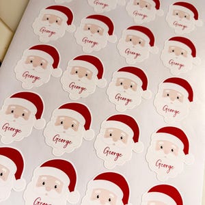 May include: A sheet of twenty individual stickers, each featuring a cartoon Santa Claus face wearing a red hat. Below each Santa face, the name George is printed in a red cursive script. The stickers are arranged in a grid pattern.