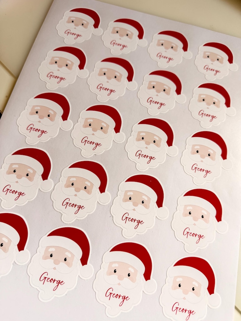 May include: A sheet of twenty individual stickers, each featuring a cartoon Santa Claus face wearing a red hat. Below each Santa face, the name George is printed in a red cursive script. The stickers are arranged in a grid pattern.