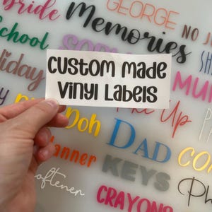 May include: A rectangular white adhesive sheet featuring the black text "custom made vinyl Labels" in a rounded sans-serif font. The sheet is held against a background displaying various colourful vinyl word decals including "TOOLS", "GEORGE", "bride", "Memories", "School", "holiday", "Doh", "DAD", "KEYS", and "CRAYONS".