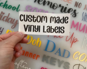 May include: A rectangular white adhesive sheet featuring the black text "custom made vinyl Labels" in a rounded sans-serif font. The sheet is held against a background displaying various colorful vinyl word decals including "TOOLS", "GEORGE", "bride", "Memories", "School", "holiday", "Doh", "DAD", "KEYS", and "CRAYONS".