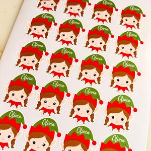 May include: A sheet of twenty-one die-cut stickers featuring a cartoon elf with brown hair in braids, wearing a red and green hat. Each sticker includes the name Olivia printed in white script on the green portion of the hat.