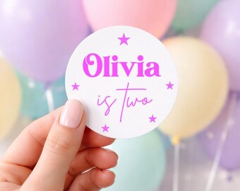 May include: A circular white sticker featuring the text Olivia is two in bright pink lettering. The design includes five small pink stars arranged in a circle around the text, set against a background of various pastel-colored balloons.