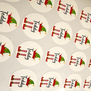 May include: A collection of circular white stickers featuring a graphic of red and white striped elf legs and a green and red elf hat. The name Teddy is printed in black script text between the legs and the hat on each individual sticker.