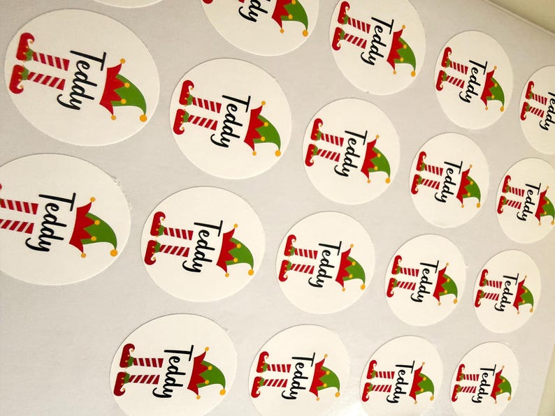 May include: A collection of circular white stickers featuring a graphic of red and white striped elf legs and a green and red elf hat. The name Teddy is printed in black script text between the legs and the hat on each individual sticker.
