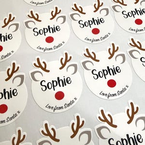 May include: A collection of circular white stickers featuring a reindeer face graphic with brown antlers, gray ears, and a red nose. Each sticker is printed with the name Sophie in black script and the text Love from Santa x below.