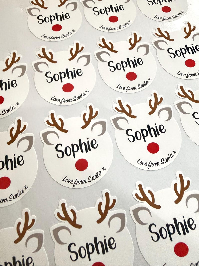 May include: A collection of circular white stickers featuring a reindeer face graphic with brown antlers, gray ears, and a red nose. Each sticker is printed with the name Sophie in black script and the text Love from Santa x below.