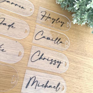 May include: A collection of clear acrylic tags with rounded ends, each featuring a small circular hole. The tags are printed with black script names including Darren, Hayley, Jade, Camilla, and Michael. The tags have a smooth, transparent finish.