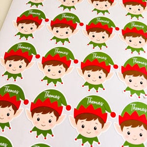 May include: A sheet of multiple circular stickers featuring a cartoon elf character with brown hair, pointed ears, and a green and red hat. Each hat displays the name Thomas in white cursive script. The stickers are arranged in a grid pattern.