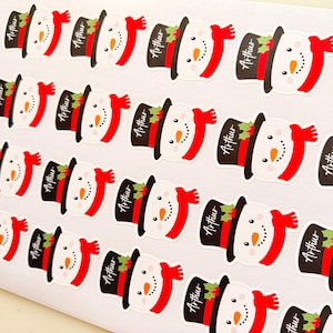May include: A sheet of multiple identical stickers featuring a cartoon snowman wearing a black top hat with a red band and green holly leaves. Each snowman has a bright orange carrot nose and a red scarf. The name Arthur is printed on each hat.