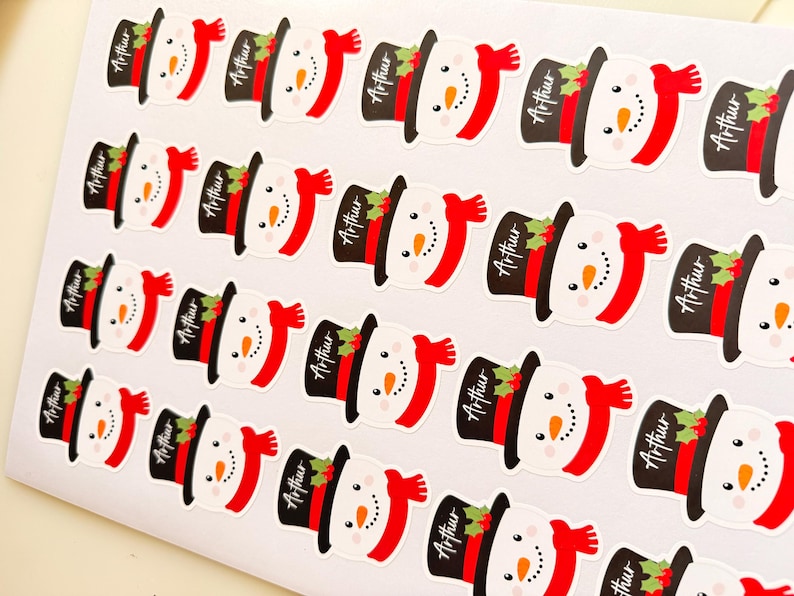 May include: A sheet of multiple identical stickers featuring a cartoon snowman wearing a black top hat with a red band and green holly leaves. Each snowman has a bright orange carrot nose and a red scarf. The name Arthur is printed on each hat.