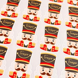 May include: A sheet of die-cut stickers featuring a cartoon nutcracker soldier with a brown mustache, wearing a red uniform and a black hat with the name Fraser written in gold script across the front of the hat brim.