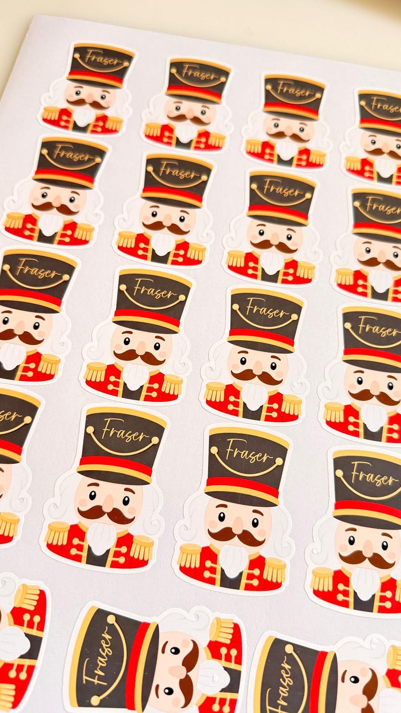 May include: A sheet of die-cut stickers featuring a cartoon nutcracker soldier with a brown mustache, wearing a red uniform and a black hat with the name Fraser written in gold script across the front of the hat brim.