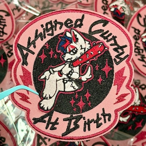 May include: Pink embroidered patch with a cartoon wolf character holding a red studded bat. The patch has the words "Affrighed Cutty At Birth" in black and red lettering, with a black background and pink stars.