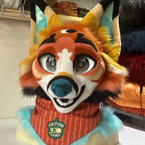 Pode incluir: Cabeça de fursuit de raposa colorida, com pelo laranja, amarelo e branco. A raposa tem olhos grandes e expressivos, um nariz azul e uma bandana vermelha com a inscrição "Critter Camp". As orelhas têm pontas pretas.