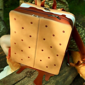 Op de afbeelding: Een rechthoekige rugzak ontworpen als een ijssandwich, met een lichtbruine buitenkant met kleine ronde inkepingen, een donkerbruine onderkant met een druipeffect en een witte tussenlaag met zilverkleurige metalen ritsen.
