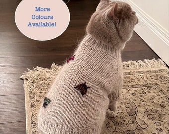 May include: A light brown cat wearing a knitted sweater with a light brown, green, and red embroidered design. The sweater has a turtleneck collar. The cat is sitting on a patterned rug. The text "More Colours Available!" is written on a pink circle in the top left corner of the image.