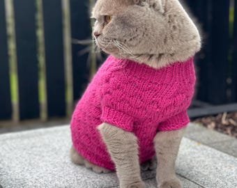 May include: A gray cat wearing a pink knitted sweater. The cat is sitting on a gray tiled surface.