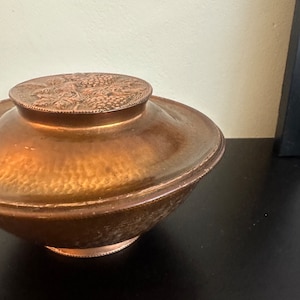 May include: A copper bowl with a lid featuring a decorative design. The bowl has a hammered finish and a raised base.
