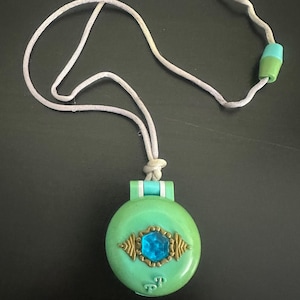Könnte beinhalten: Eine grüne und blaue Halskette mit einem runden Anhänger, der einen blauen Edelstein mit goldenen Akzenten zeigt. Der Anhänger hat die Buchstaben "TP" auf der Unterseite.
