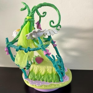 Può includere: Una statuina stravagante con una struttura di vite verde con fiori bianchi a forma di margherita e accenti rosa. Un tessuto verde chiaro drappeggia la vite e la base è un ovale verde chiaro e viola. La statuina ha un tema fiabesco.