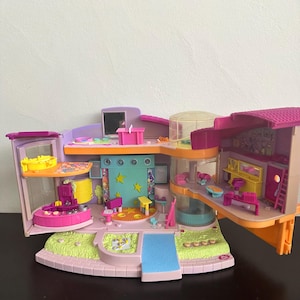 Op de afbeelding: Een kleurrijk poppenhuis met meerdere niveaus, met verschillende kamers en functies. Het poppenhuis heeft een roze, paars en geel kleurenschema, met een zwembad, een podium en meubels. Het poppenhuis is ontworpen voor fantasierijk spel.