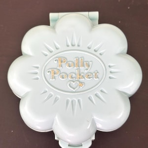 Può includere: Un compatto Polly Pocket azzurro a forma di fiore. Il compatto ha un coperchio incernierato e presenta il logo Polly Pocket in lettere dorate. I petali del fiore hanno bordi appuntiti.