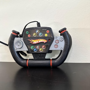 Op de afbeelding: Een zwart en grijs Hot Wheels racestuur met een getextureerde grip, rode accenten en een middenconsole met meerdere kleurrijke ronde knoppen, een richtingspad en een bedrade aansluitkabel die aan de bovenkant uit het apparaat komt.