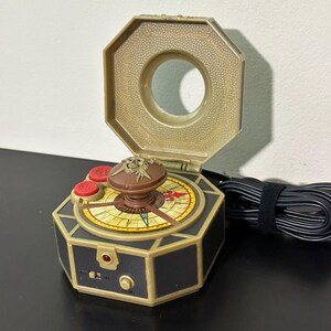 以下が含まれることがあります： 金と黒のおもちゃのコンパス、ヒンジ付きの蓋付き。 コンパスは、中央のノブ、赤いボタン、装飾的な羅針盤のデザインを備えた詳細な文字盤を備えています。 黒いコードが側面に付いています。