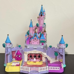 Può includere: Un castello giocattolo in plastica a più livelli con un'alta torre centrale rosa, tetti conici blu, pareti viola e dettagli dorati. L'interno comprende una zona pranzo con tavolo, sedie, una scala e un orologio sulla facciata.