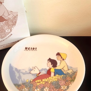Könnte beinhalten: Weiße Keramikplatte mit einer farbenfrohen Illustration von Heidi, einem Mädchen aus den Alpen, mit einem Jungen und einem Hund, umgeben von Blumen und Bergen. Auf der Platte steht "HEIDI A GIRL OF THE ALPS".