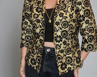 Pode incluir: Blazer com padrão floral dourado e preto com mangas três quartos. O casaco tem lapelas clássicas e um corte sob medida. O design floral é um padrão repetido de flores estilizadas. O blazer é usado com um top preto e jeans de lavagem escura.