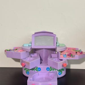 Peut inclure: Un ensemble de jeu de coiffeuse en plastique lavande comprenant un miroir central, plusieurs tiroirs coulissants et des compartiments latéraux. L'extérieur est orné de vignes vertes peintes et de fleurs bleues et roses, avec des boutons de tiroir roses.