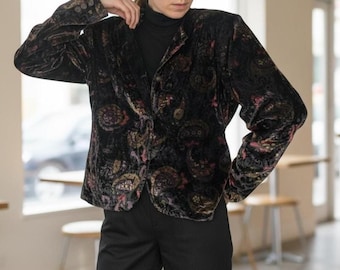 May include: Black velvet blazer with a paisley pattern in shades of gold, pink, and brown. The blazer has a classic collar and button closure. The model is wearing a black turtleneck and black pants.