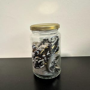May include: A clear glass jar with a gold-coloured metal lid filled with a collection of assorted jewellery pieces, including silver-toned mesh bracelets, dark beaded strands, and various metallic chains visible through the transparent glass walls.