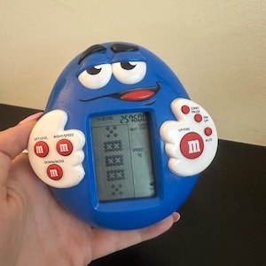 Op de afbeelding: Een blauw elektronisch handheld spel met M&M's-thema. Het spel heeft een digitaal scherm met een Tetris-achtig spel, met knoppen met de tekst "Left/Level", "Right/Speed" en "Down/Mode". Het spel heeft een highscore van 259500.