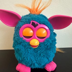 Puede incluir: Un juguete Furby azul turquesa con orejas rosas, pies rosas y pelo naranja. El Furby tiene grandes ojos naranjas y un pico amarillo.