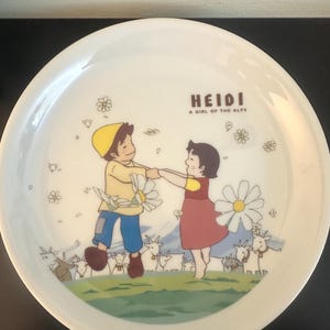 Könnte beinhalten: Weißer Keramikteller mit einer farbenfrohen Illustration von zwei tanzenden Kindern, umgeben von Ziegen und Blumen. Der Teller trägt den Text "HEIDI A GIRL OF THE ALPS" in braunen Buchstaben.