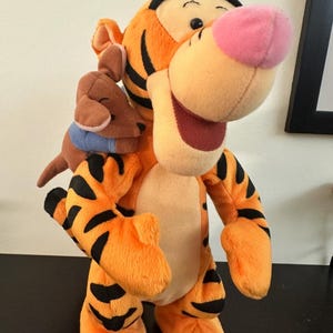 Könnte beinhalten: Ein Plüschtier von Tigger, einem orangefarbenen Tiger mit schwarzen Streifen, und einem kleinen braunen Känguru. Tigger hat eine rosa Nase und ein großes Lächeln. Das Känguru trägt einen blauen Kragen. Die Spielzeuge stehen auf einer schwarzen Oberfläche.