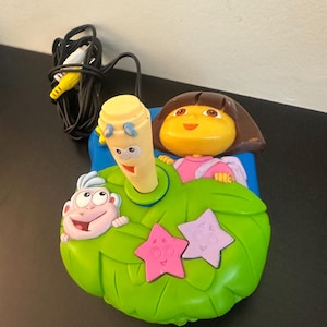 Puede incluir: Un juguete infantil con los personajes de Dora la Exploradora y Botas. El juguete tiene una base verde en forma de hoja con detalles de estrellas rosas y moradas. También se ve un micrófono amarillo y un cable negro con conectores amarillos y blancos.