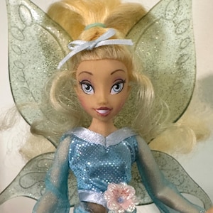 Puede incluir: Muñeca de hada rubia con el pelo recogido en una coleta con un lazo azul claro. La muñeca viste un top azul con lentejuelas y una falda a juego. Tiene grandes alas brillantes y un adorno de flor rosa en la cintura.