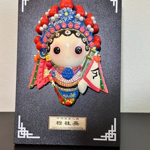 Peut inclure: Figurine d'opéra chinois colorée, montée sur une plaque noire avec des coins blancs décoratifs. La figurine porte une coiffe détaillée avec des perles rouges et un motif floral bleu. Elle tient un petit drapeau.