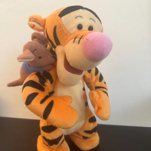 Könnte beinhalten: Ein Plüschtier von Tigger, einem orangefarbenen Tiger mit schwarzen Streifen, einer rosa Nase und einem lächelnden Gesichtsausdruck. Ein kleines braunes Känguru-Spielzeug ist an Tiggers Rücken befestigt. Das Spielzeug steht auf einer schwarzen Oberfläche.