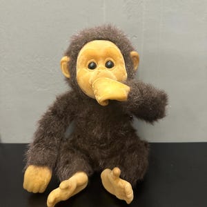 May include: A plush chimpanzee toy with dark brown fur and a light tan face, ears, hands, and feet. The chimpanzee has black eyes and is posed with one hand raised towards its mouth. The toy features a soft, textured body and smooth vinyl-like limbs.