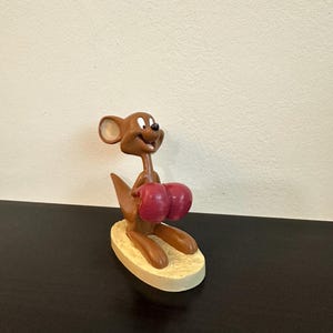 Puede incluir: Una figura de ratón de dibujos animados marrón con guantes de boxeo rojos. El ratón está posado sobre una base ovalada beige. La figura tiene una expresión sonriente y está sobre un fondo neutro.