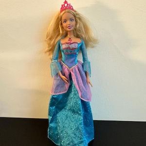 Puede incluir: Una muñeca princesa con pelo largo rubio, ojos azules y una tiara rosa. Lleva un vestido largo color turquesa con un corpiño y mangas rosas y moradas. El vestido tiene una falda estampada y brillante.